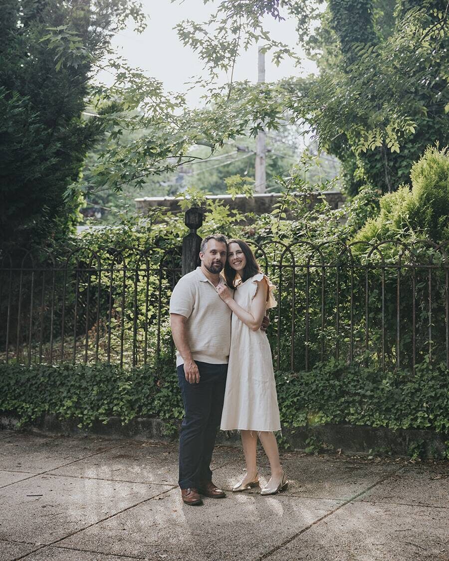 Ben and Shannon in front of an ironwork garden fence, under summer trees