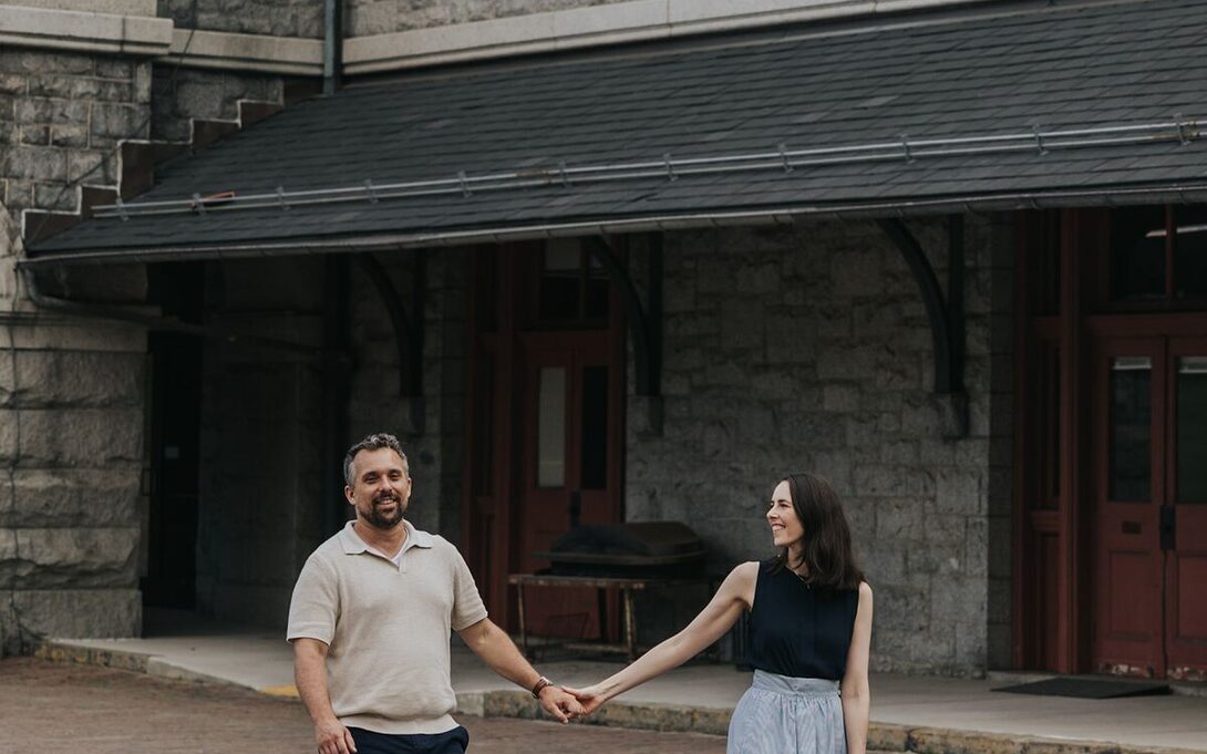 Ben and Shannon walking hand in hand past a stone building in Baltimore